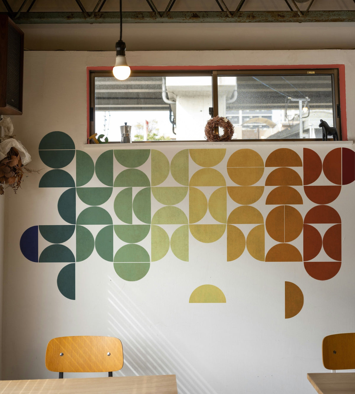 A large geometric wall mural with a retro pixel-art style design in a gradient of blue, green, yellow, and orange, displayed on a white wall in a cafe interior with wooden tables and chairs.